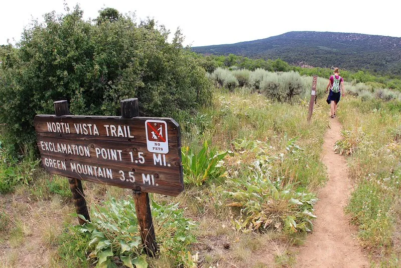 A wooden sign marks the North Vista Trail toward Exclamation Point and Green Mountain as a hiker walks along a narrow dirt path through shrubs and open terrain.