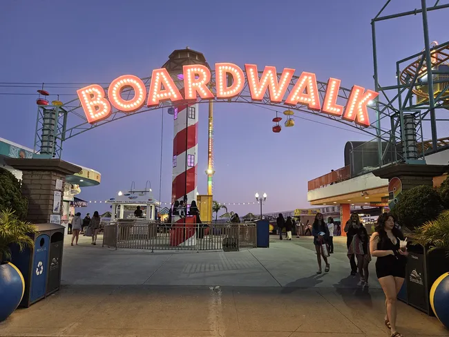 A brightly lit “Boardwalk” sign arches over the entrance to a seaside amusement park at dusk, with colorful lights glowing against the evening sky. Visitors stroll beneath the sign among rides, snack stands, and a striped lighthouse-themed attraction in the center of the plaza.
