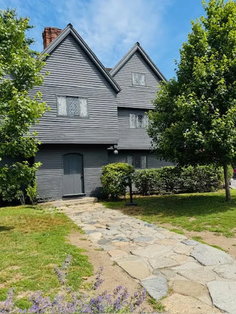 A historic dark gray wooden house with diamond-paned windows and pitched gables, framed by green trees and a stone path leading to the entrance.