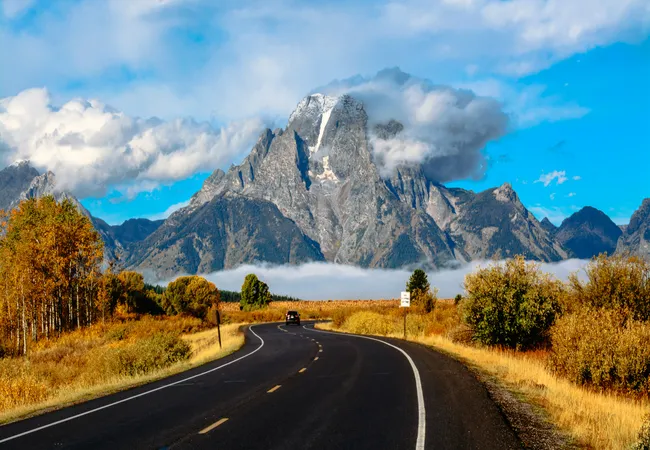 winding two-lane asphalt road leads toward a massive, snow-dusted mountain peak partially covered by a thick white cloud. The surrounding landscape features yellow and orange autumn foliage under a bright blue sky.