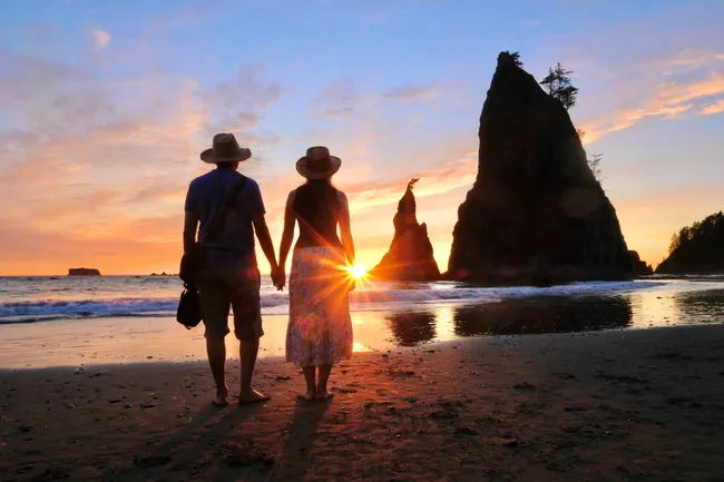 A couple stands hand in hand on a beach at sunset, with dramatic rock formations and gentle waves in the background at Olympic National Park.