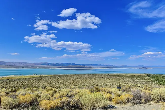 Expansive lake surrounded by dry grassy terrain and distant mountains under a bright blue sky with scattered clouds.