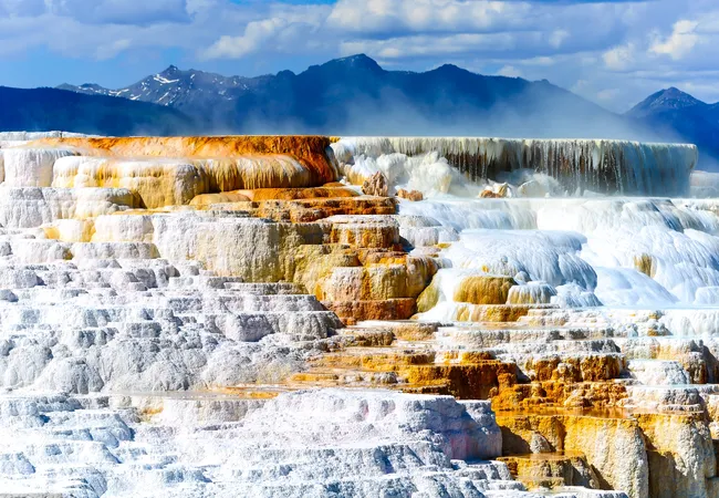 A large, multi-tiered natural limestone terrace is covered in flowing water, creating a series of white and bright orange mineral pools. Wisps of white steam rise from the upper levels against a backdrop of distant blue mountains and a bright sky with scattered clouds.
