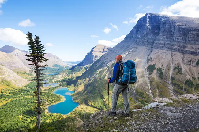 Hiker with backpack stands on a cliff overlooking turquoise lakes and dramatic mountain peaks in Glacier National Park.