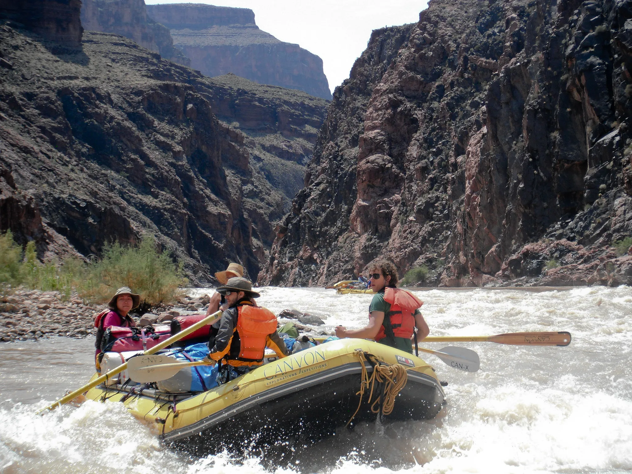A group of people wearing life jackets white-water rafting on a yellow inflatable boat through the churning rapids of the Colorado River at the bottom of the Grand Canyon.