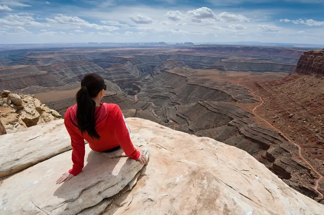 Woman sitting on a cliff edge overlooking the deep winding canyons of Muley Point.