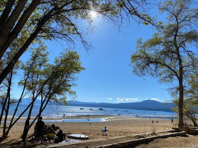 Sunny lakeside beach with people relaxing and boats on the water, framed by trees and distant mountains.