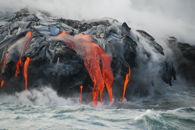 Molten lava from Kīlauea Volcano flows into the ocean, creating steam as it meets the crashing waves.