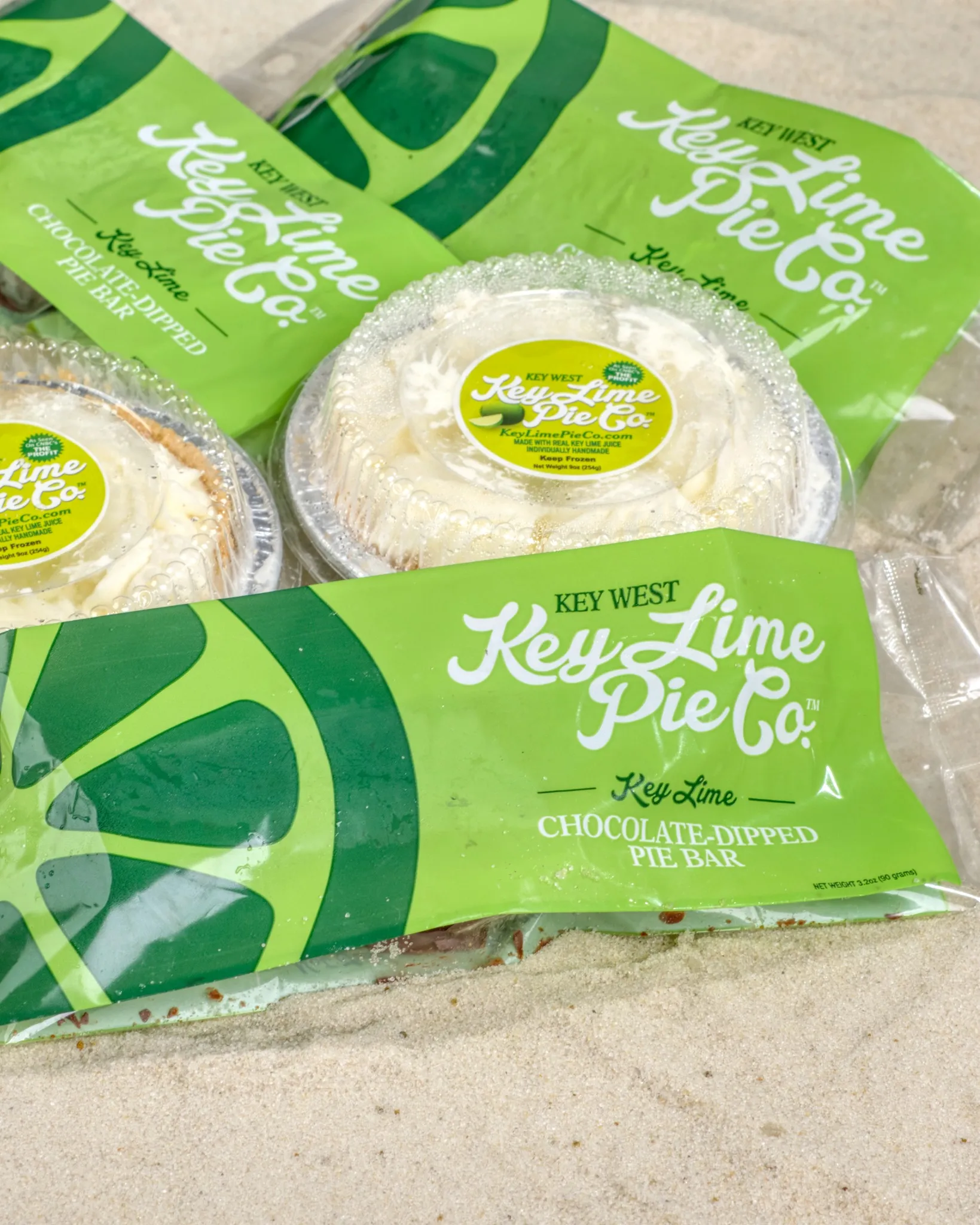 A flat-lay, close-up shot of several packaged desserts from "Key West Key Lime Pie Co." resting on light, fine-grained sand. The image features bright green plastic wrappers for "Chocolate-Dipped Pie Bars" and two small, individual key lime pies in clear plastic domed containers with yellow and green labels. The branding is bold and vibrant against the neutral sand background