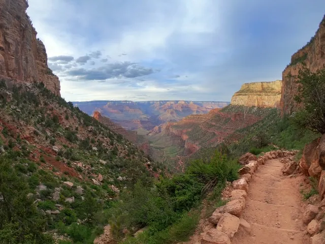 A dusty, winding trail hugs the canyon wall, bordered by rough stones, overlooking a vast landscape of layered red cliffs, green valleys, and a hazy blue horizon under a soft, cloud-streaked sky.