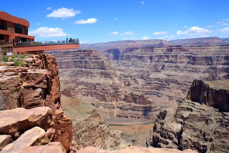A wide, daytime landscape of the Grand Canyon Skywalk. On the left, a large, horseshoe-shaped glass bridge juts out over a deep, vertical red rock cliff. Small figures of people can be seen walking on the bridge. The massive canyon depth reveals layered brown and orange rock faces, with the Colorado River visible far below as a muddy brown line. The scene is set under a bright blue sky with scattered white clouds.