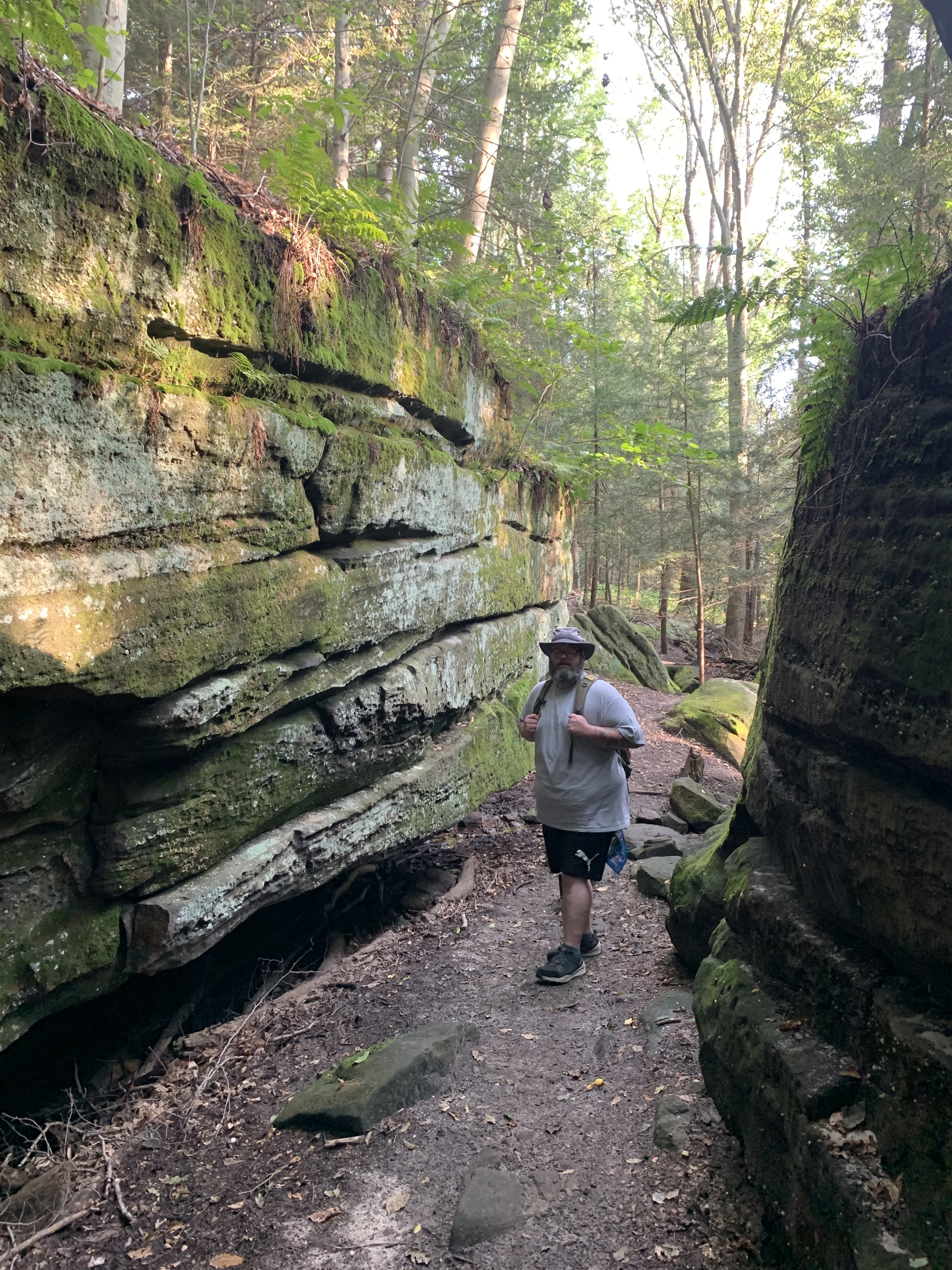A hiker wearing a gray t-shirt, black shorts, a bucket hat, and a backpack walks along a narrow dirt trail between towering moss-covered sandstone ledges on the Ledges Trail in Cuyahoga Valley National Park, Ohio. The layered rock walls rise steeply on both sides of the path, draped in green moss, lichen, and ferns. Tall trees filter soft dappled light from above, creating an atmospheric, canyon-like forest passage.