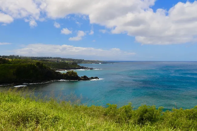 Scenic coastal view of Maui with turquoise water, rocky cliffs, and lush green hills under a bright, partly cloudy sky.