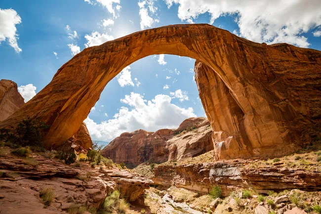 A stunning low-angle landscape shot of Rainbow Bridge, a massive natural sandstone arch. The bridge forms a perfect orange-brown arc across a deep canyon, with jagged red rock formations in the background. The sun shines from behind the top of the arch, creating a bright glow against a vibrant blue sky filled with fluffy white clouds. The rugged canyon floor in the foreground features layers of weathered rock and sparse green vegetation.