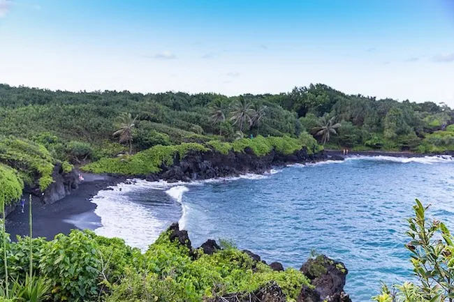 A secluded black sand beach with turquoise waves, lava rock shoreline, and lush tropical greenery along the coast.