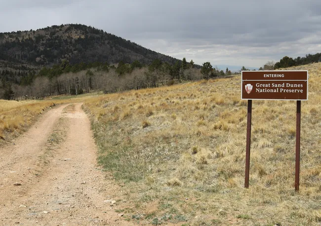 A dirt trail leads into the grassy highlands of Great Sand Dunes National Preserve, marked by a brown entrance sign with forested hills and overcast skies in the background.